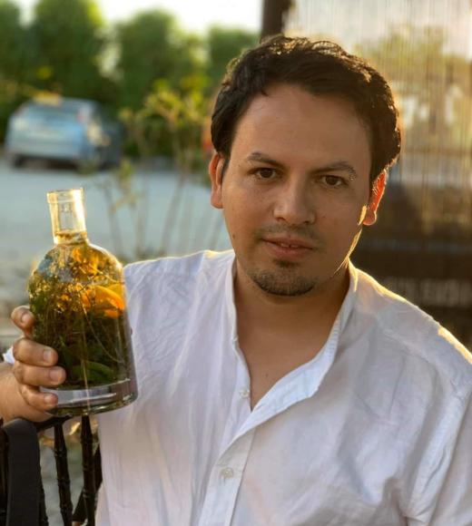 Carlos Barahona in a white shirt holiding a bottle filled with herbs and green liquid