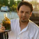 Carlos Barahona in a white shirt holiding a bottle filled with herbs and green liquid